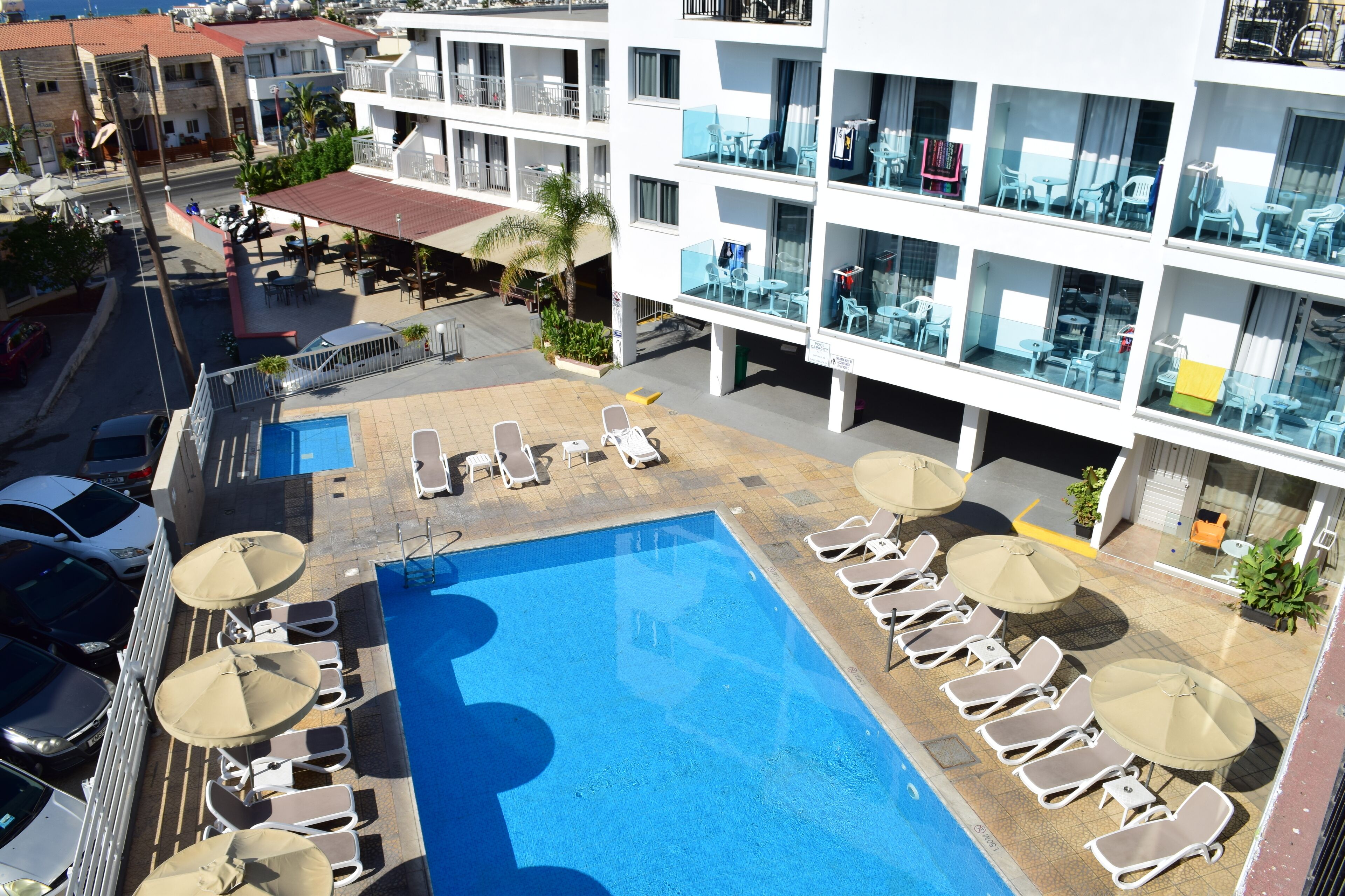 Un área de piscina en la azotea con sillones de descanso y sombrillas, rodeada por un edificio de varios pisos con balcones y zonas de estar al aire libre.
