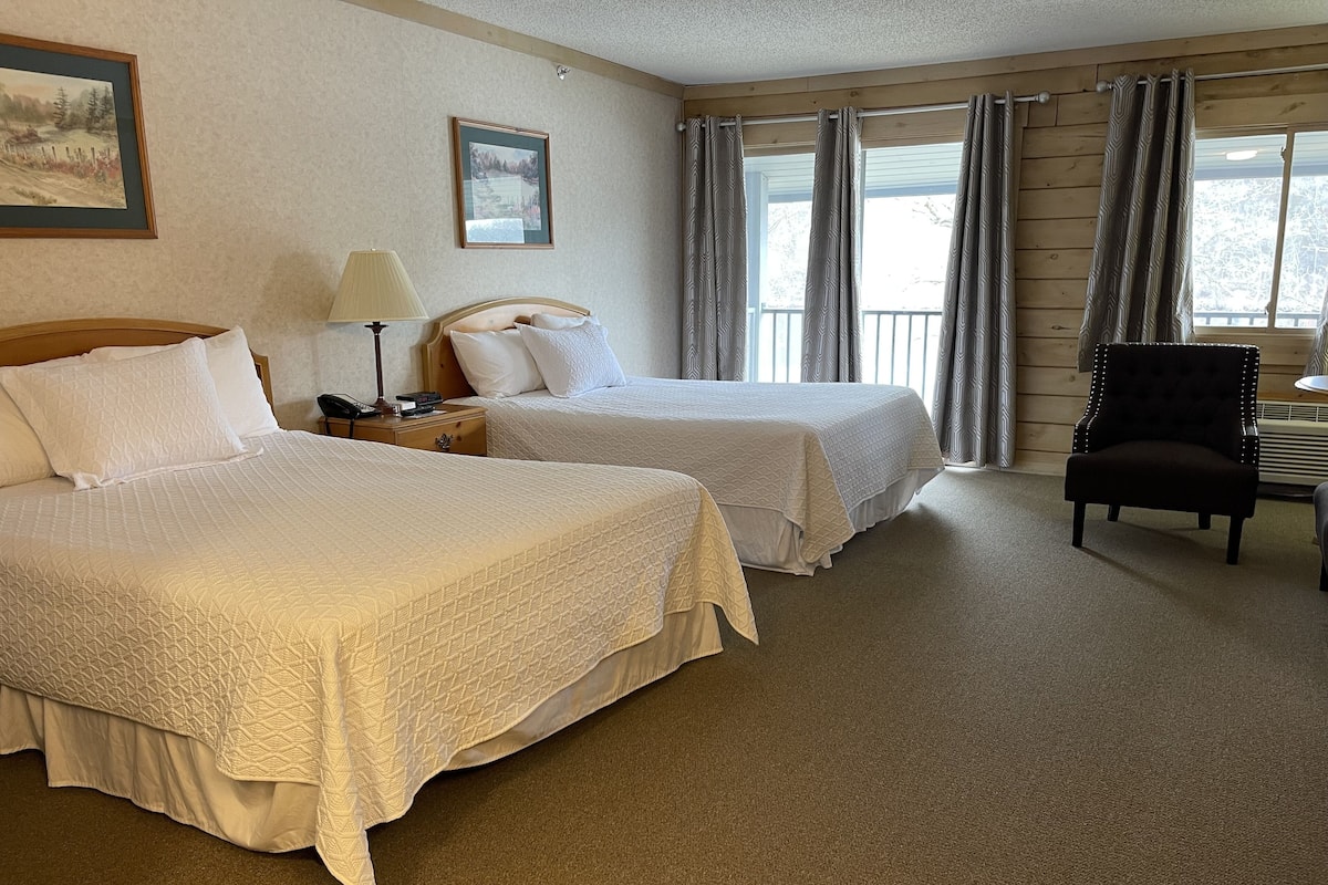 A hotel room with two beds, a desk, a chair, and large windows with curtains.