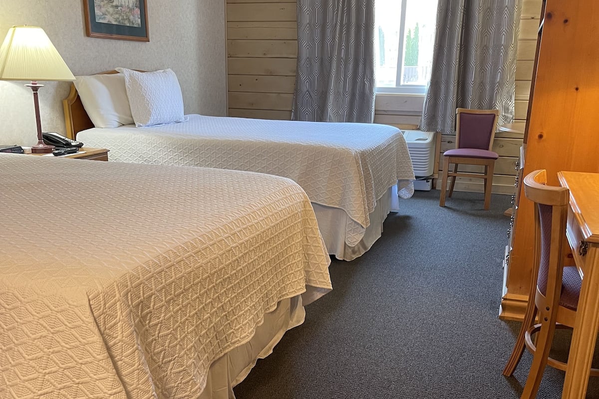 A hotel room with two beds, a desk, a chair, a lamp, and a window with curtains.