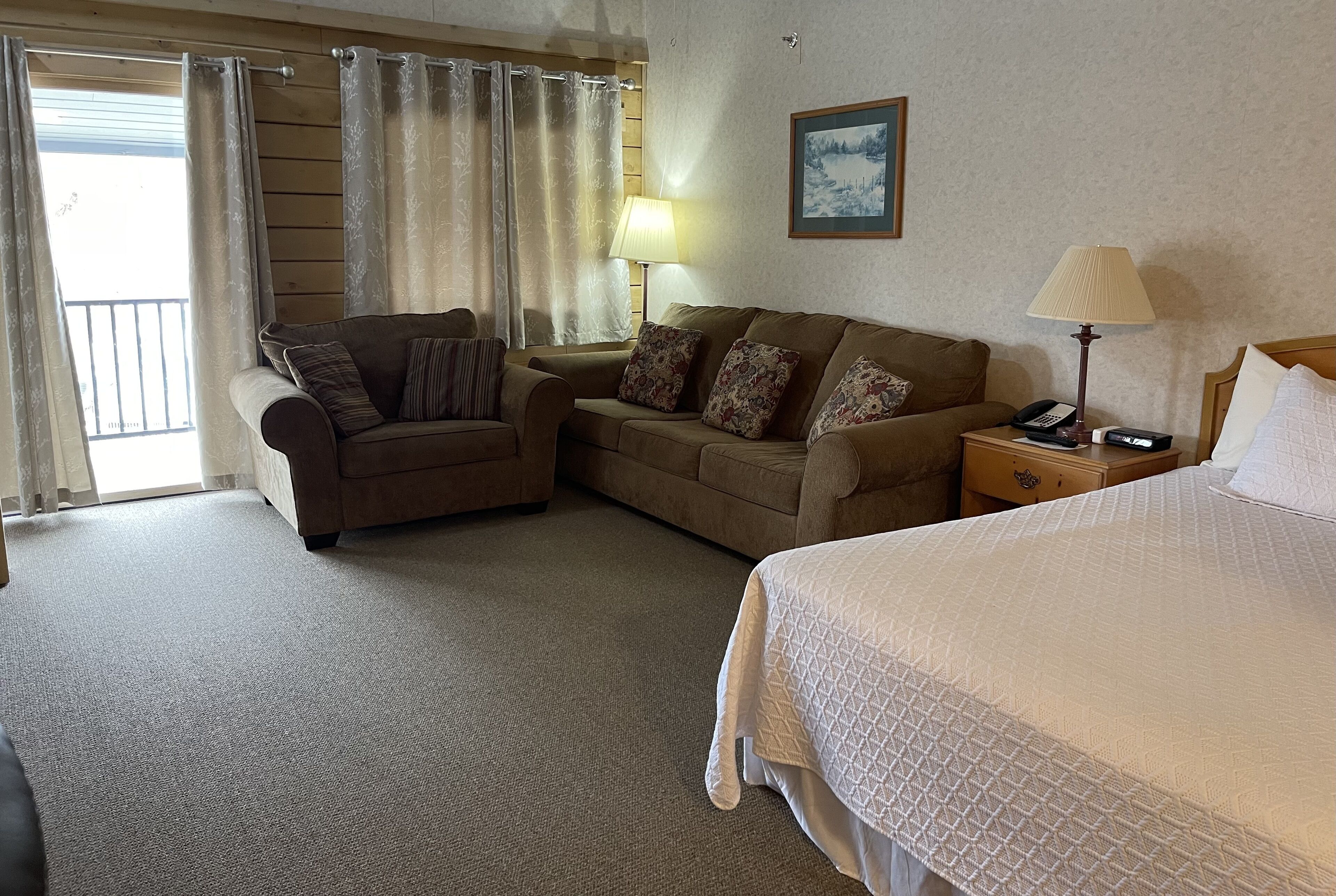 Habitación de hotel con cama, dos sofás, una mesita de noche con lámpara, un cuadro en la pared y vistas al exterior a través de la ventana.