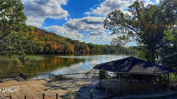 A lakeside gazebo with a wooden deck and railing, overlooking a calm lake and autumn foliage.