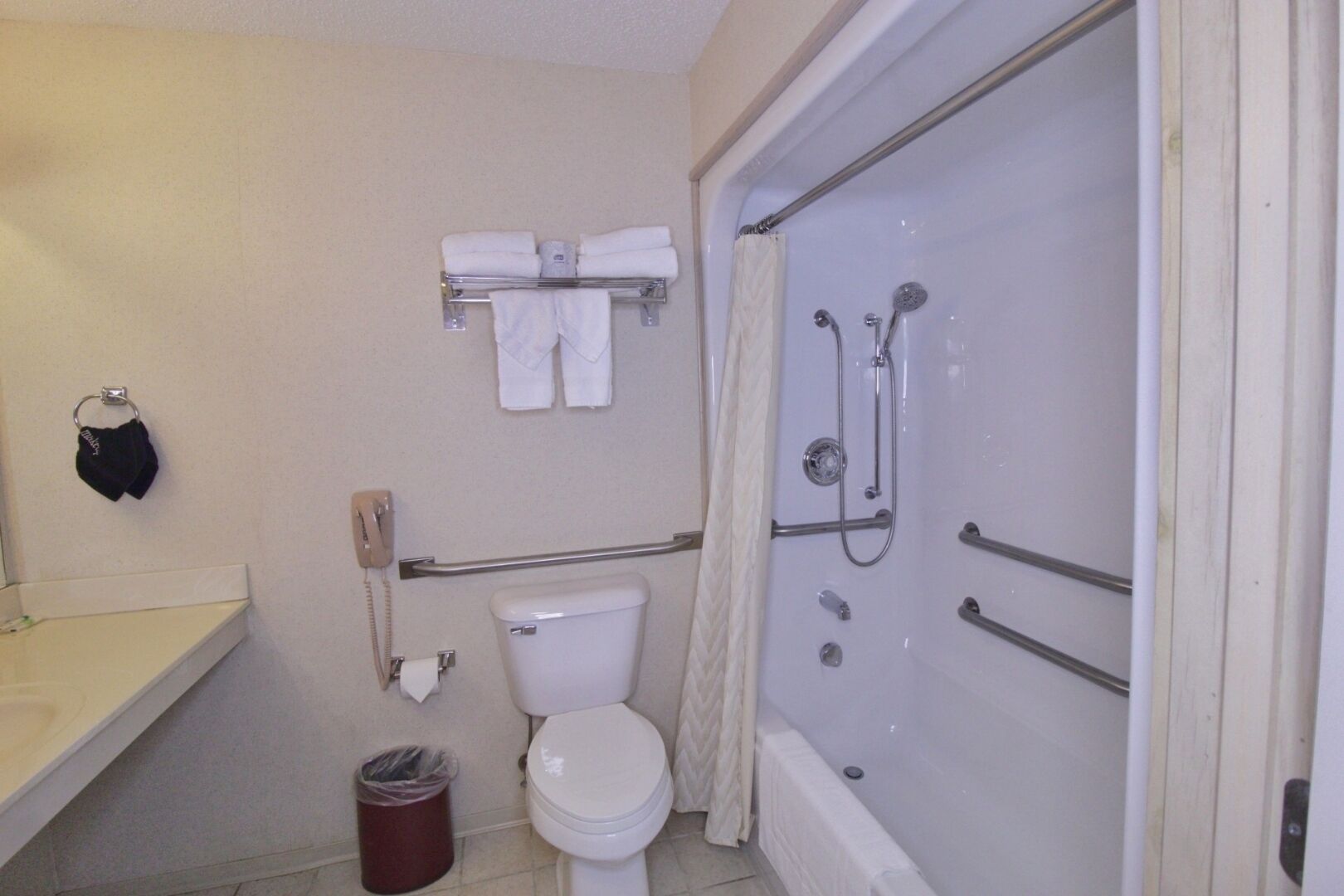 Un baño con inodoro, ducha con barras de apoyo, toallero con toallas, cesto de basura y teléfono.