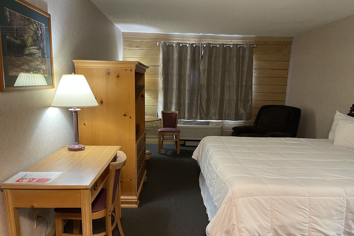 A hotel room with a bed, a desk with a lamp, a chair, a wardrobe, and a window with curtains.