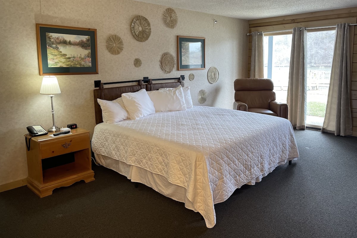 A hotel room with a large bed, a bedside table with a lamp, a chair, and a view of the outdoors through the window.