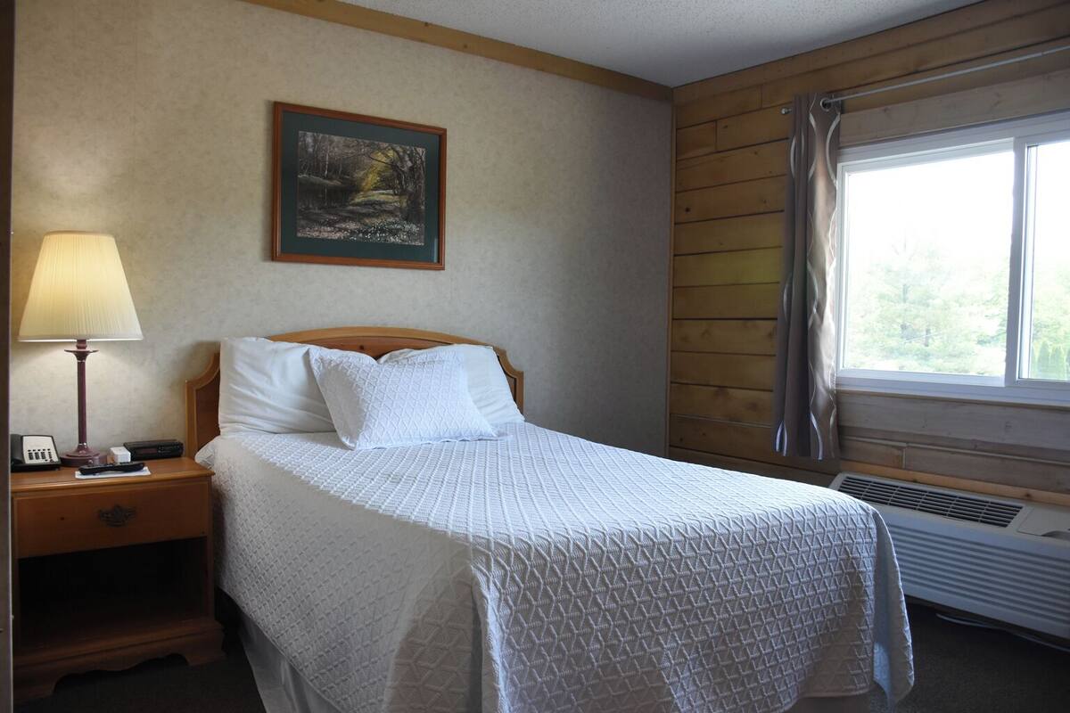 A neatly made bed with a quilted white bedspread, a wooden headboard, a bedside lamp, a telephone, and a framed picture on the wall.
