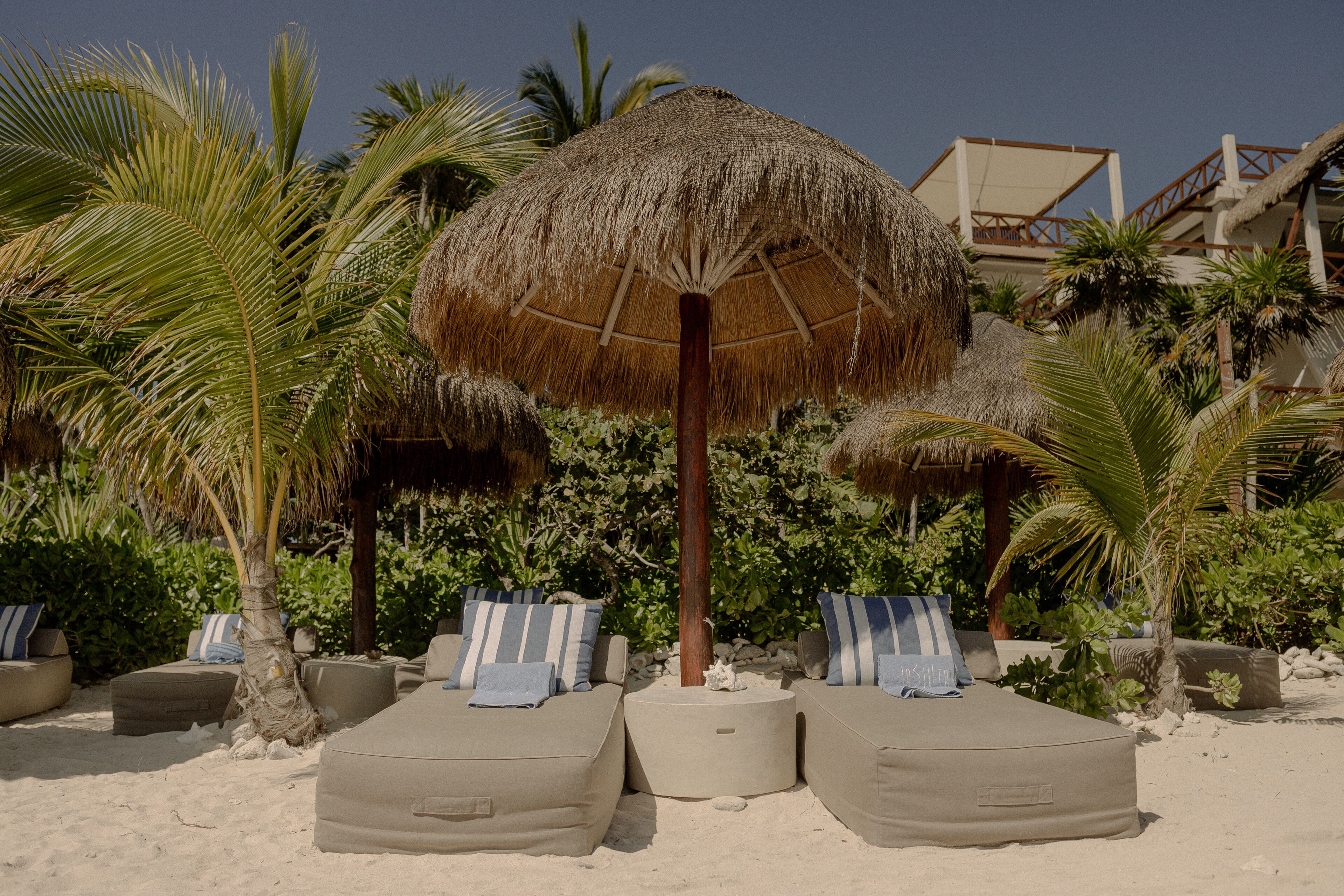 Un área de descanso en la playa con sombrillas de paja, asientos acolchados y palmeras.