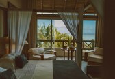 A bedroom with a large bed, two chairs, a small table, and a view of the ocean through large windows.