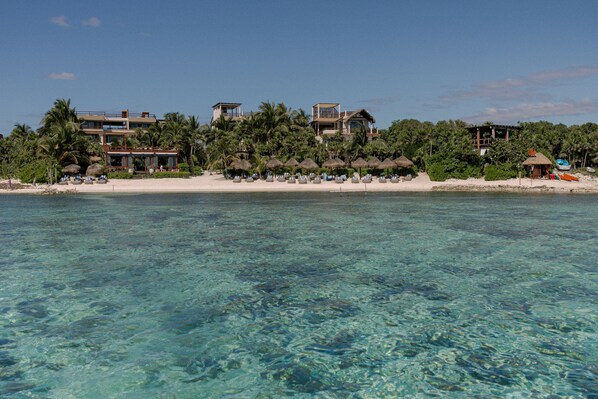 Una propiedad frente a la playa con varias edificaciones, agua turquesa cristalina y una playa de arena con sombrillas.