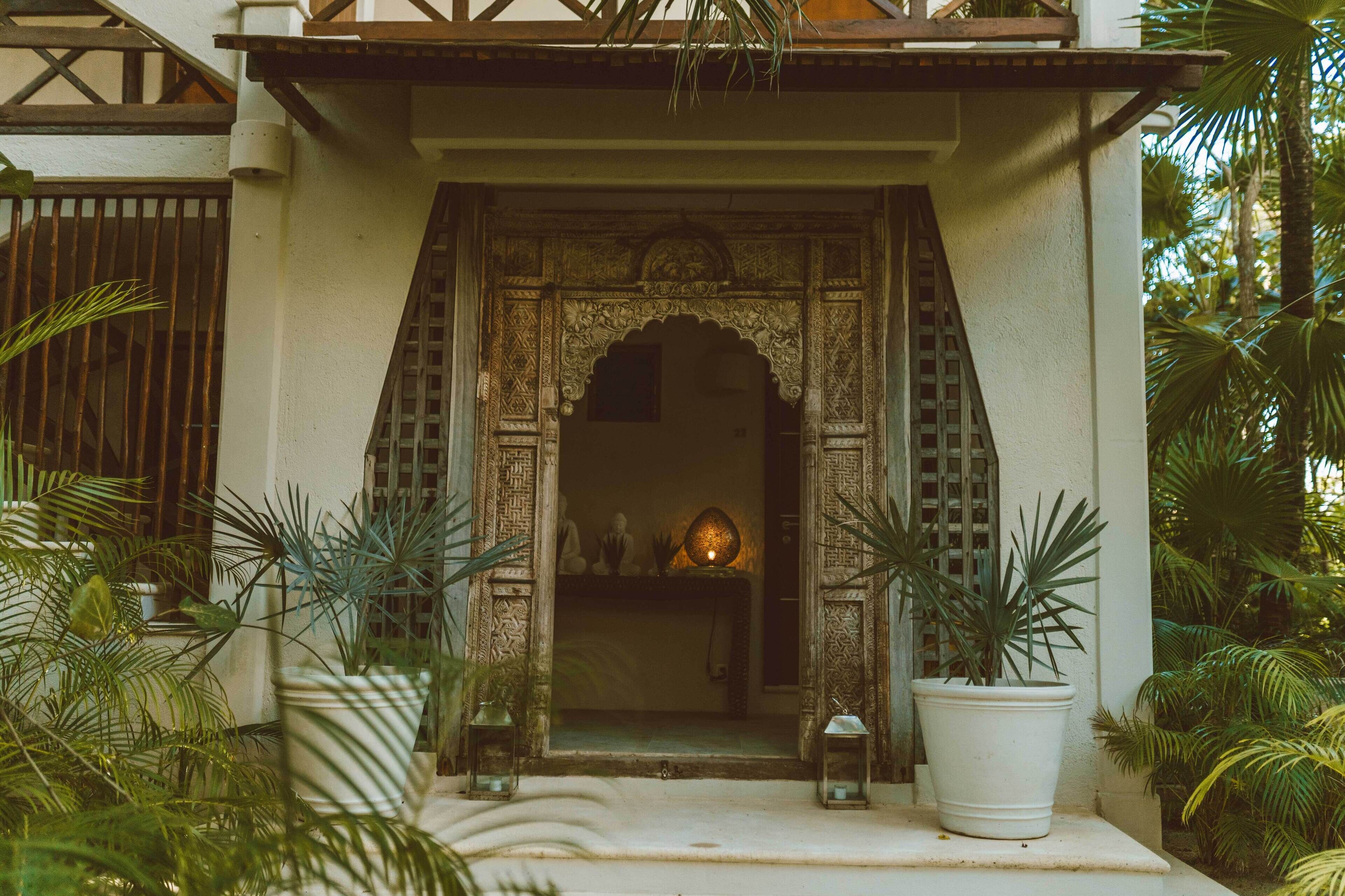A traditional entrance with intricate carvings, a small shelf with decorative items, and potted plants.