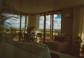A hotel room with a large balcony, a bed, a sofa, a table with chairs, and a lamp.