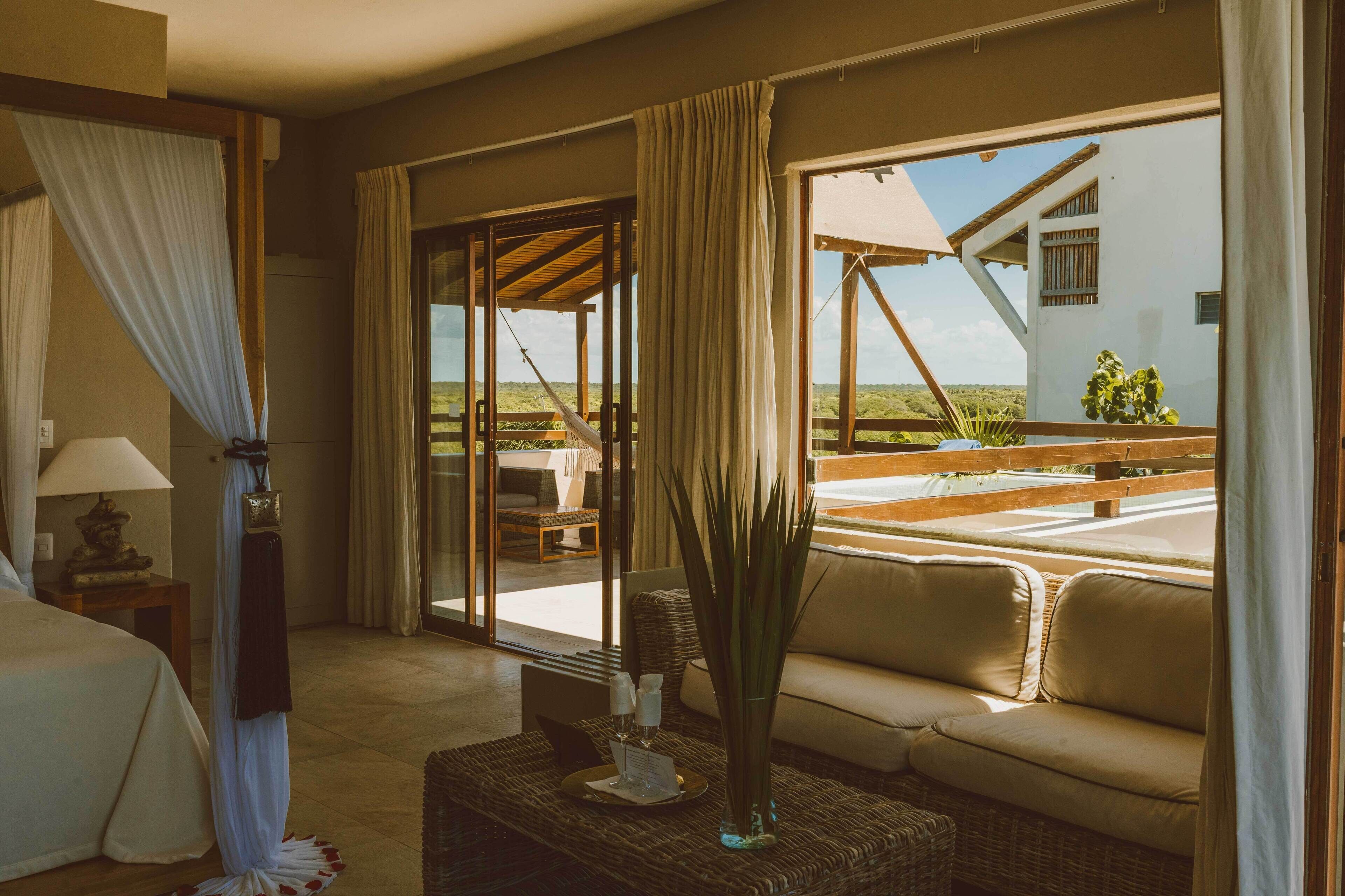 A hotel room with a bed, a sofa, a table with a vase, and large windows offering a view of the outdoors.