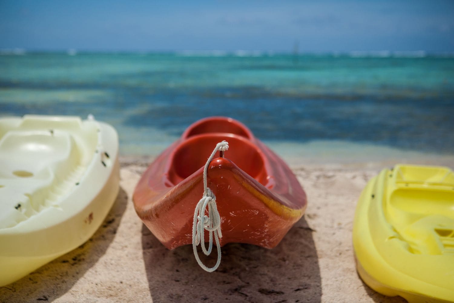 Three colorful kayaks on a sandy beach with a clear blue ocean and a distant ship in the background.