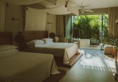 A hotel room with two beds, a ceiling fan, a large window with a view of greenery, and a balcony with a chair.