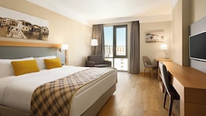 Une chambre d’hôtel comprenant un lit, deux chaises, un bureau, une télévision et un balcon avec des rideaux.