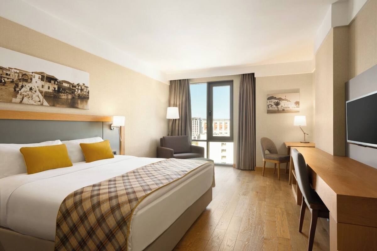 Een hotelkamer met een bed, twee stoelen, een bureau, een televisie en een balkon met gordijnen.