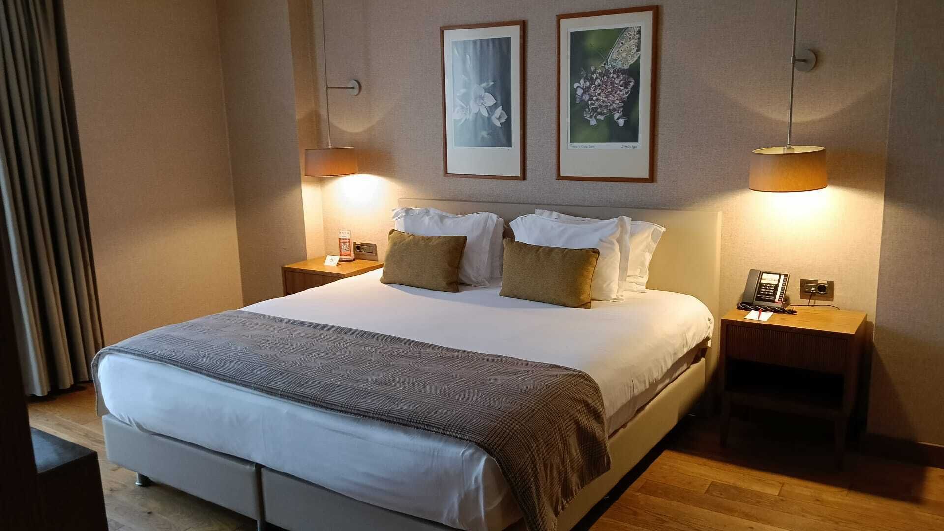 A hotel room with a large bed, two bedside tables, a lamp, and two framed pictures on the wall.