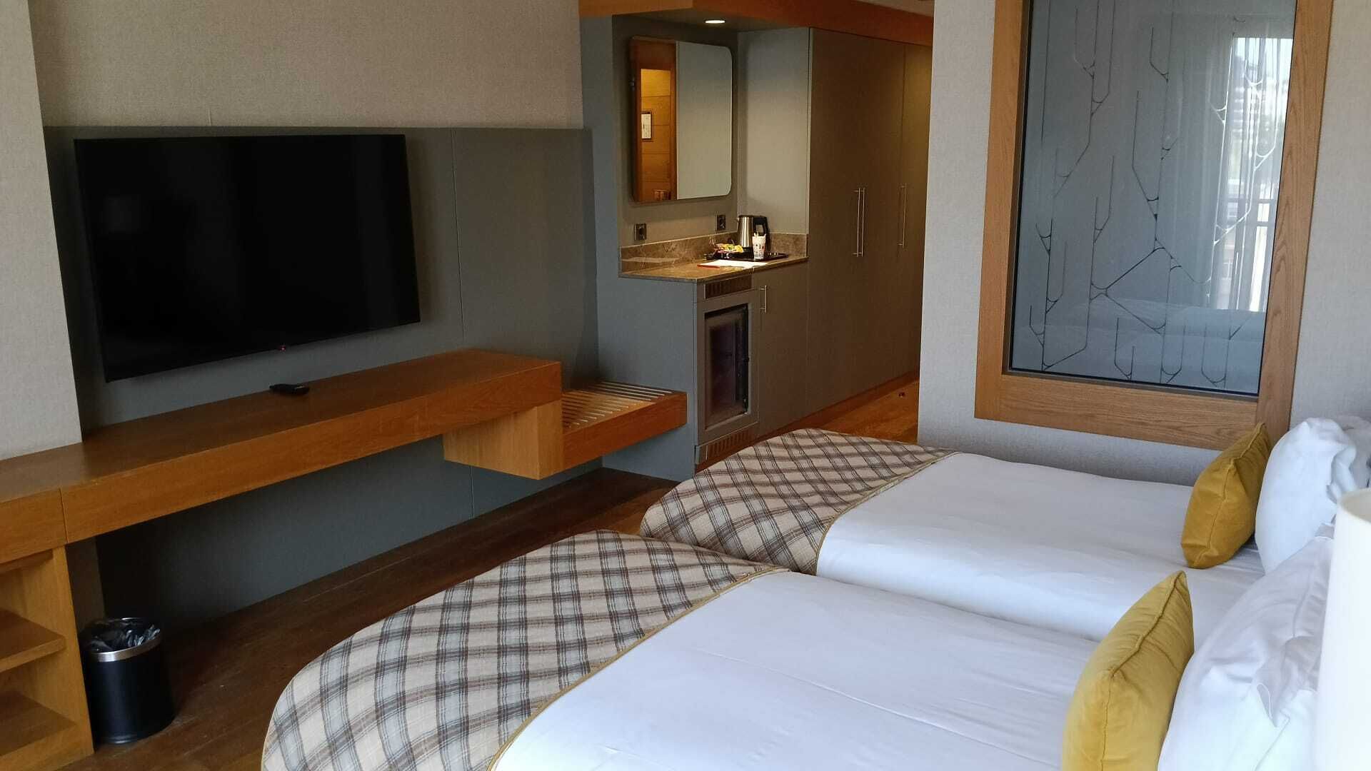 A hotel room with a TV, a wooden bench, a bed with plaid bedding, a sink, and a window with a view.