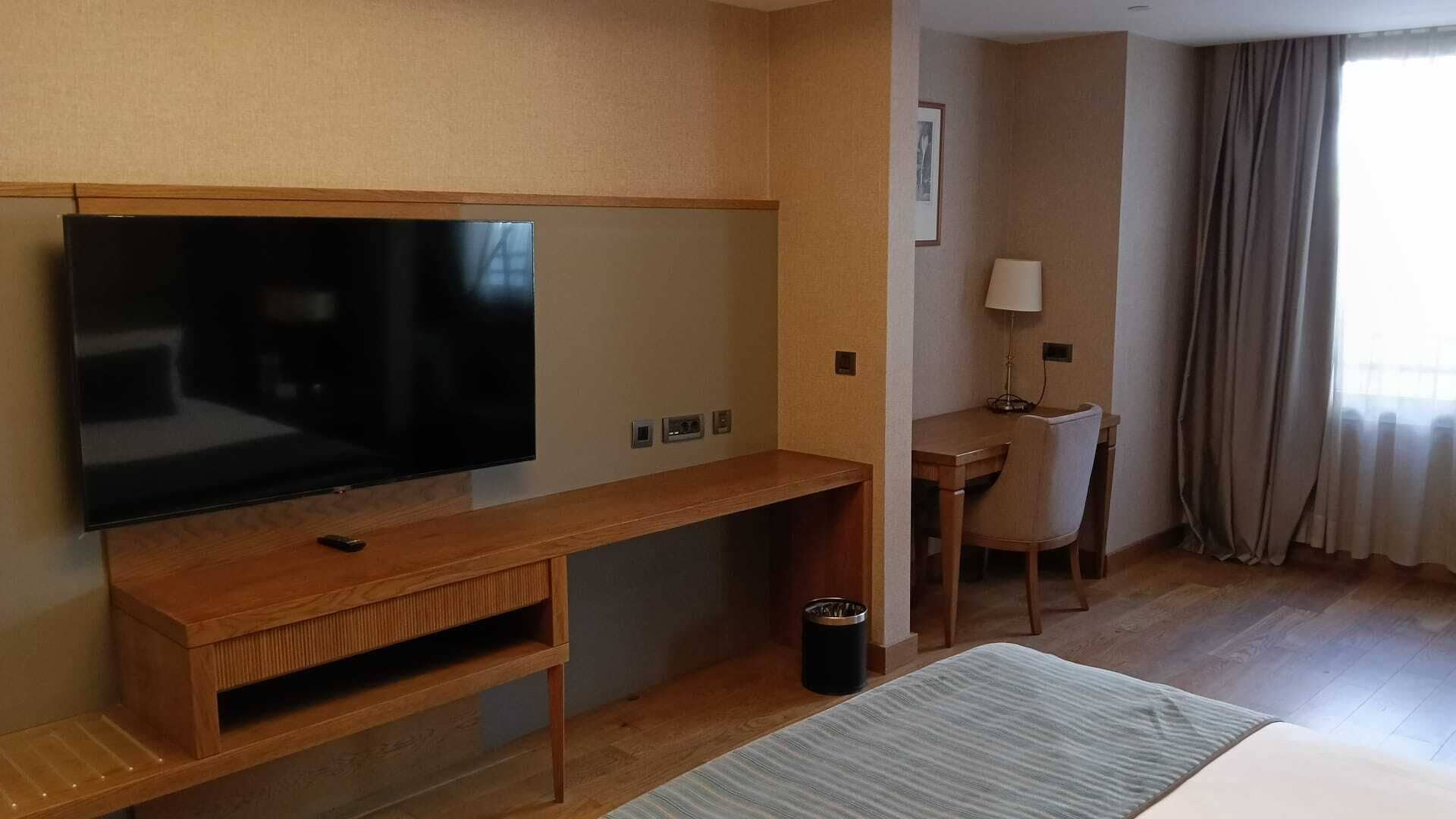 Une chambre d’hôtel équipée d’un téléviseur à écran plat fixé sur une console en bois, d’un bureau avec une chaise, d’une lampe et d’une fenêtre avec des rideaux.