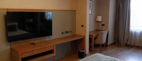 Une chambre d’hôtel équipée d’un téléviseur à écran plat fixé sur une console en bois, d’un bureau avec une chaise, d’une lampe et d’une fenêtre avec des rideaux.