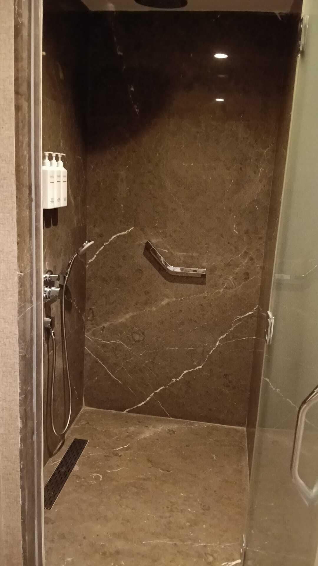 Chuveiro com parede de mármore, chuveiro manual e um frasco de shampoo.