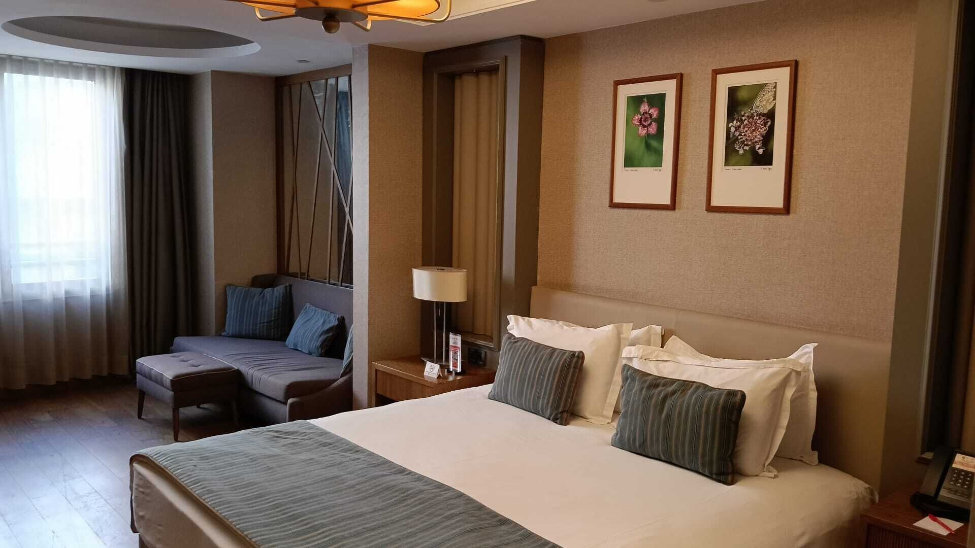 A hotel room with a large bed, a sofa, a desk with a lamp, and two framed pictures on the wall.