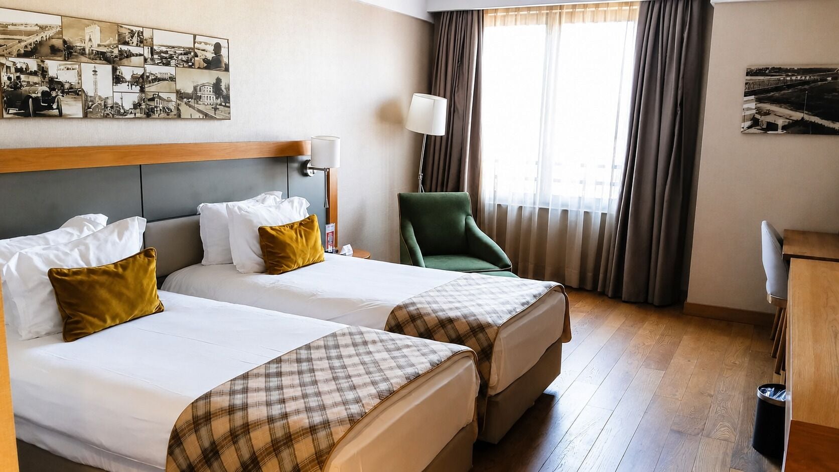 A hotel room with two beds, a green armchair, a desk, and a large window with curtains.