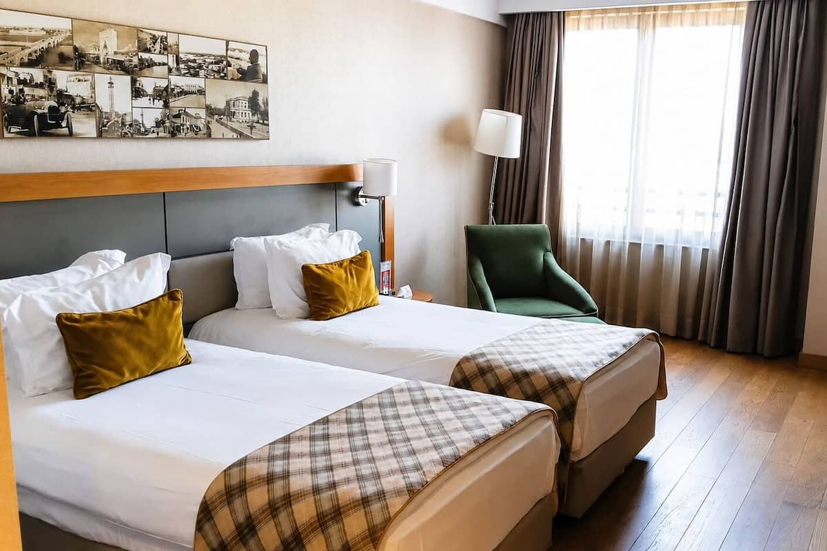 Een hotelkamer met twee bedden, een groene fauteuil, een bureau en een groot raam met gordijnen.