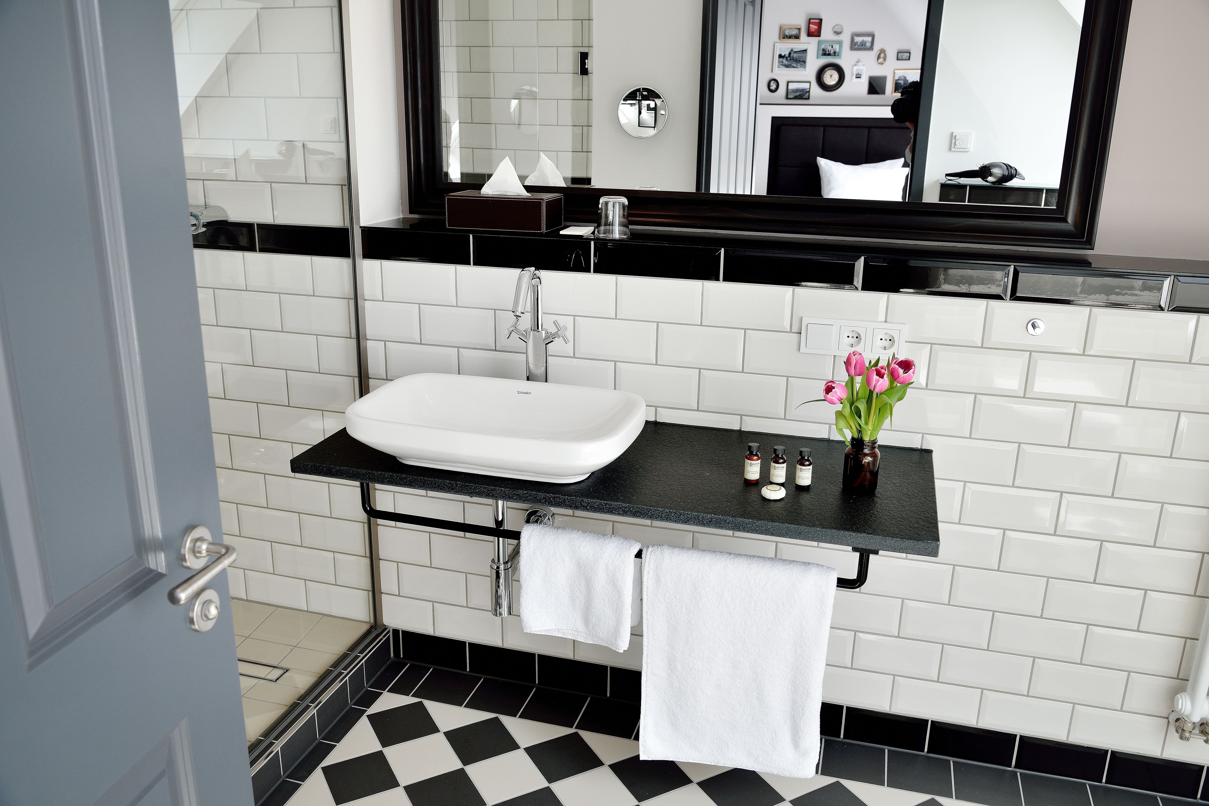 Ein Badezimmer mit weißem Waschbecken, schwarzer Arbeitsplatte und einem Spiegel darüber. Ein Handtuch hängt an der Wand und auf der Arbeitsplatte steht ein kleiner Blumenstrauß.