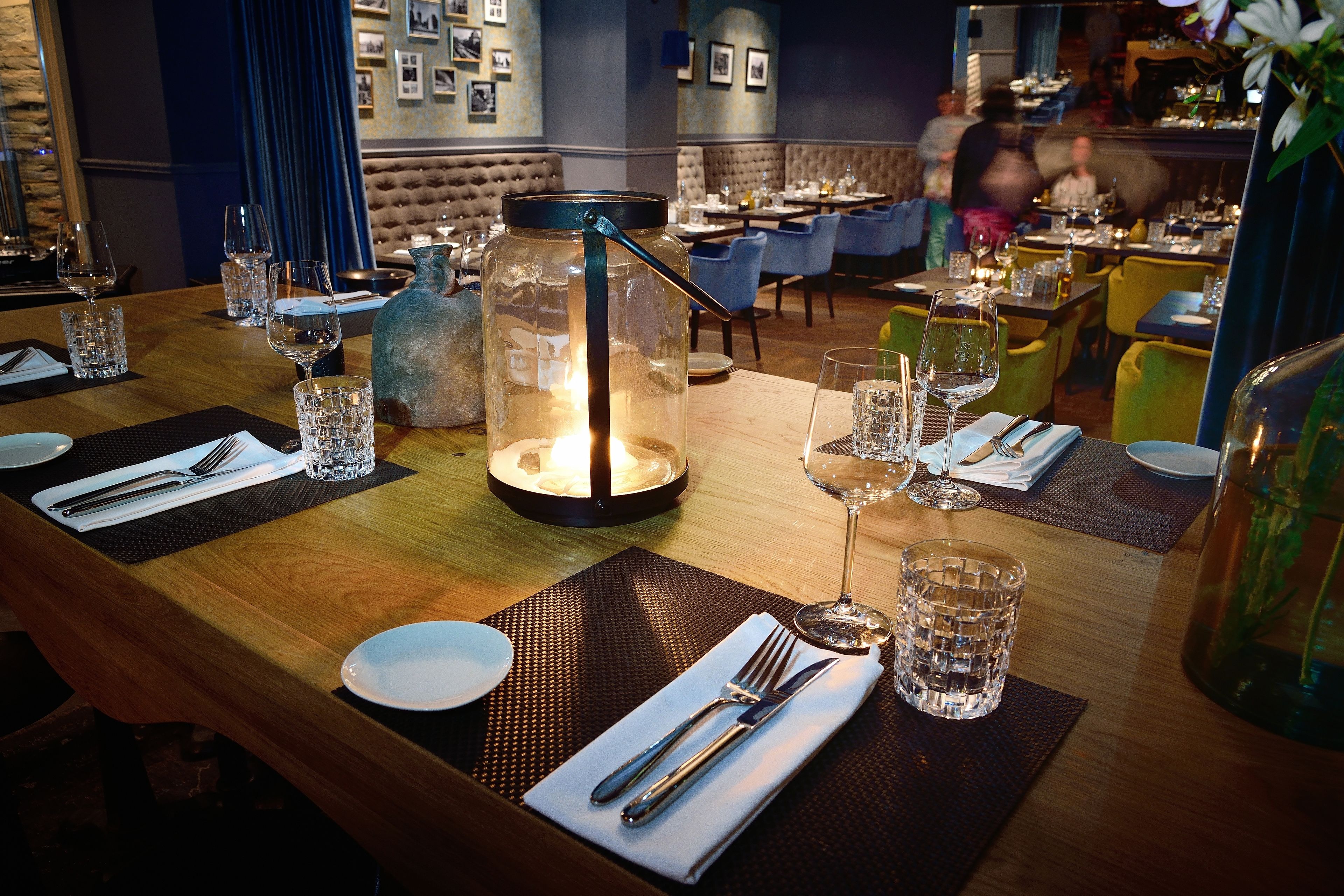 Ein Restaurantinneres mit gedeckten Tischen, einem dekorativen Glasarrangement in der Mitte und einer blauen, gepolsterten Sitzbank im Hintergrund.