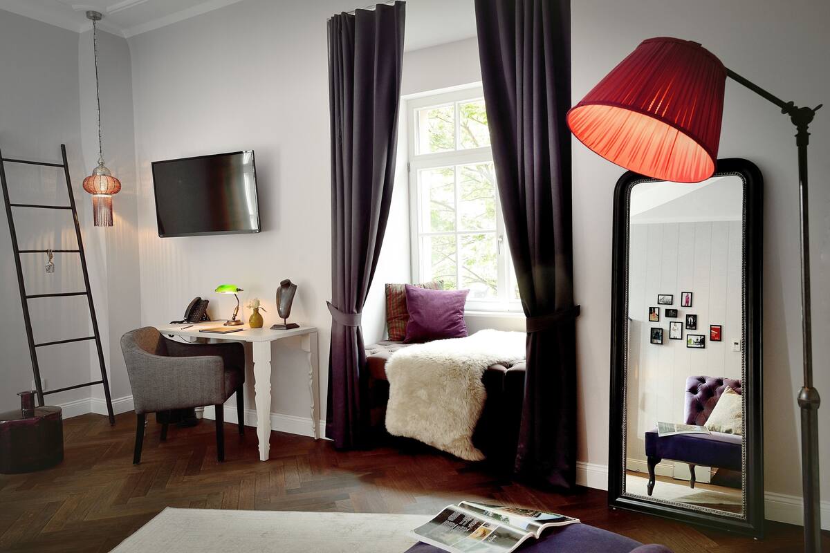 A bedroom with a bed, a desk, a chair, a mirror, and a window with curtains.