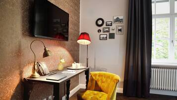 Ein Zimmer mit Wandfernseher, Schreibtisch mit Lampe, Stuhl und blickdichten Vorhängen am Fenster.