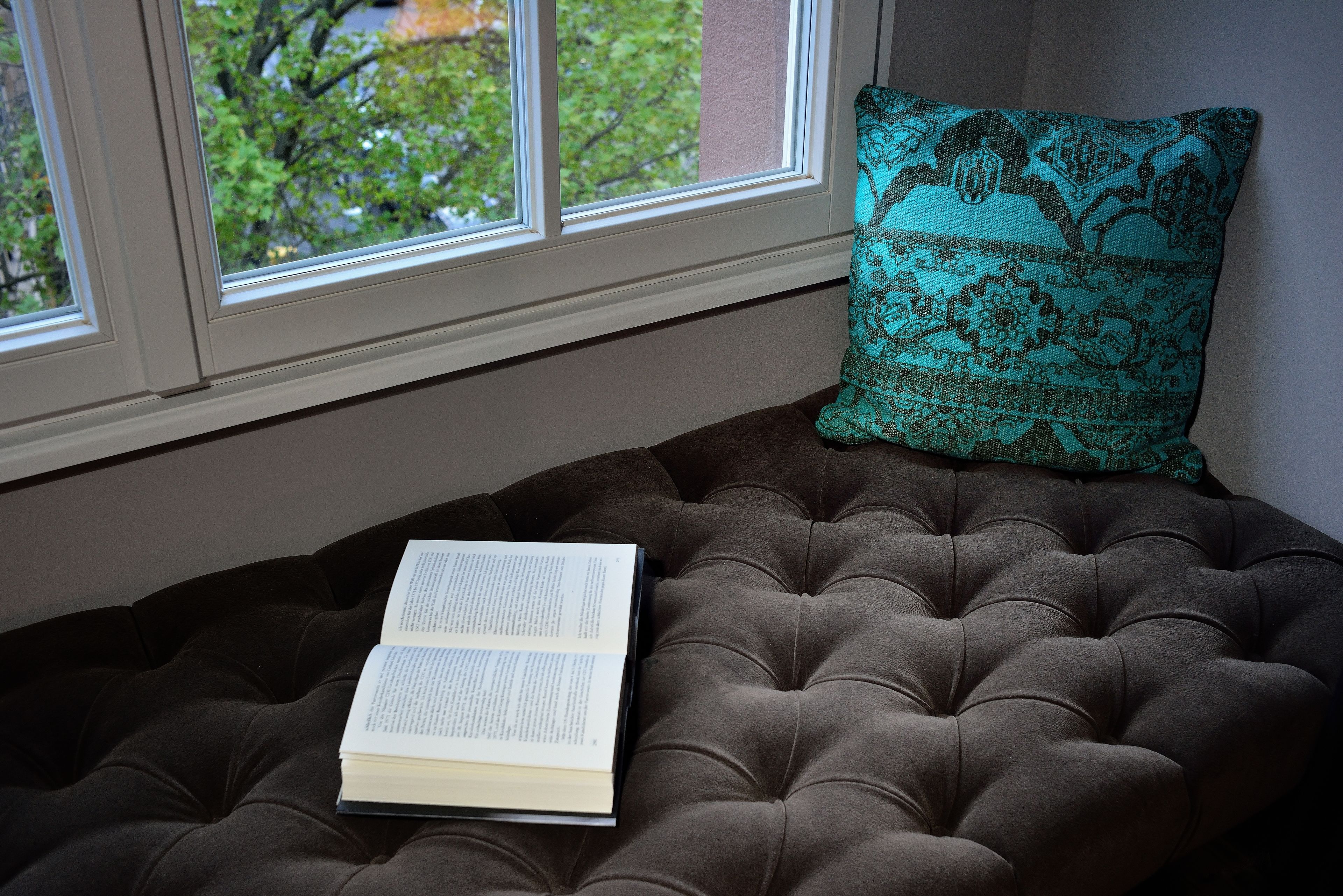 Un livre posé sur un canapé à accoudoirs capitonnés, près d’une fenêtre, avec un coussin à motifs.