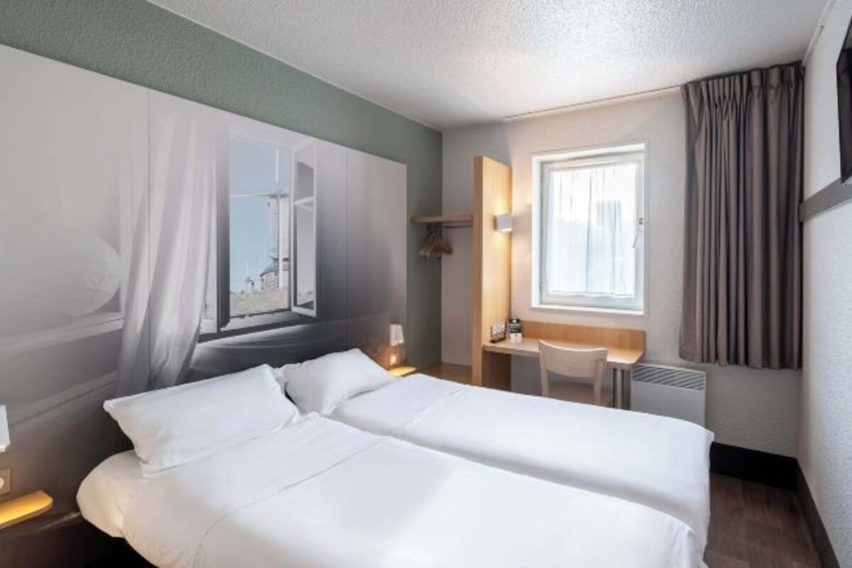 A hotel room with two beds, a desk, a chair, a window with curtains, and a wall-mounted light.