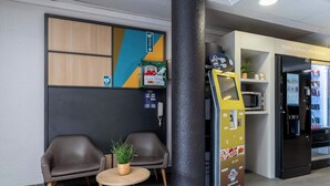 Een moderne wachtruimte met twee leren fauteuils, een tafeltje met een plant, een pilaar, een automaat en een koelkast met een menu.