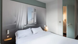 Een hotelkamer met een groot bed, een nachtkastje met een lamp, een raam met dunne gordijnen en een deur naar een badkamer.