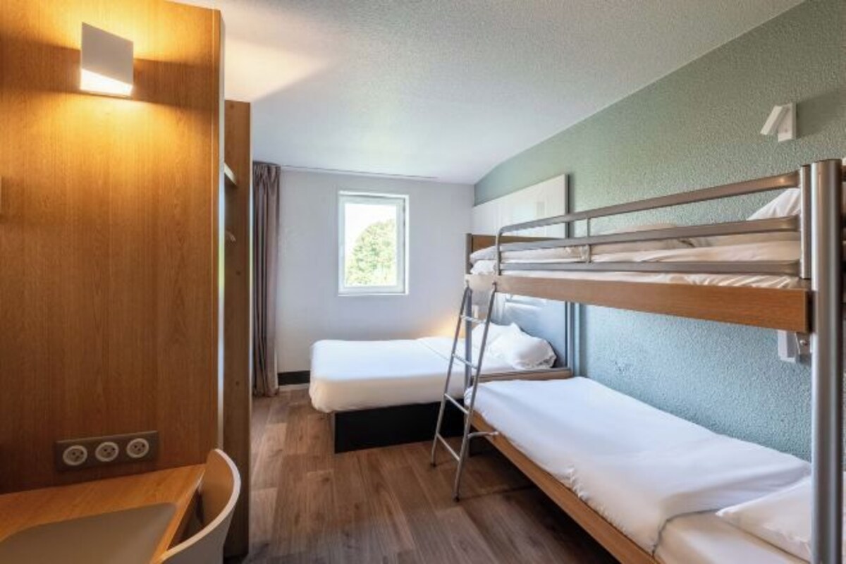 A compact hotel room with bunk beds, a small desk, and a window providing natural light.