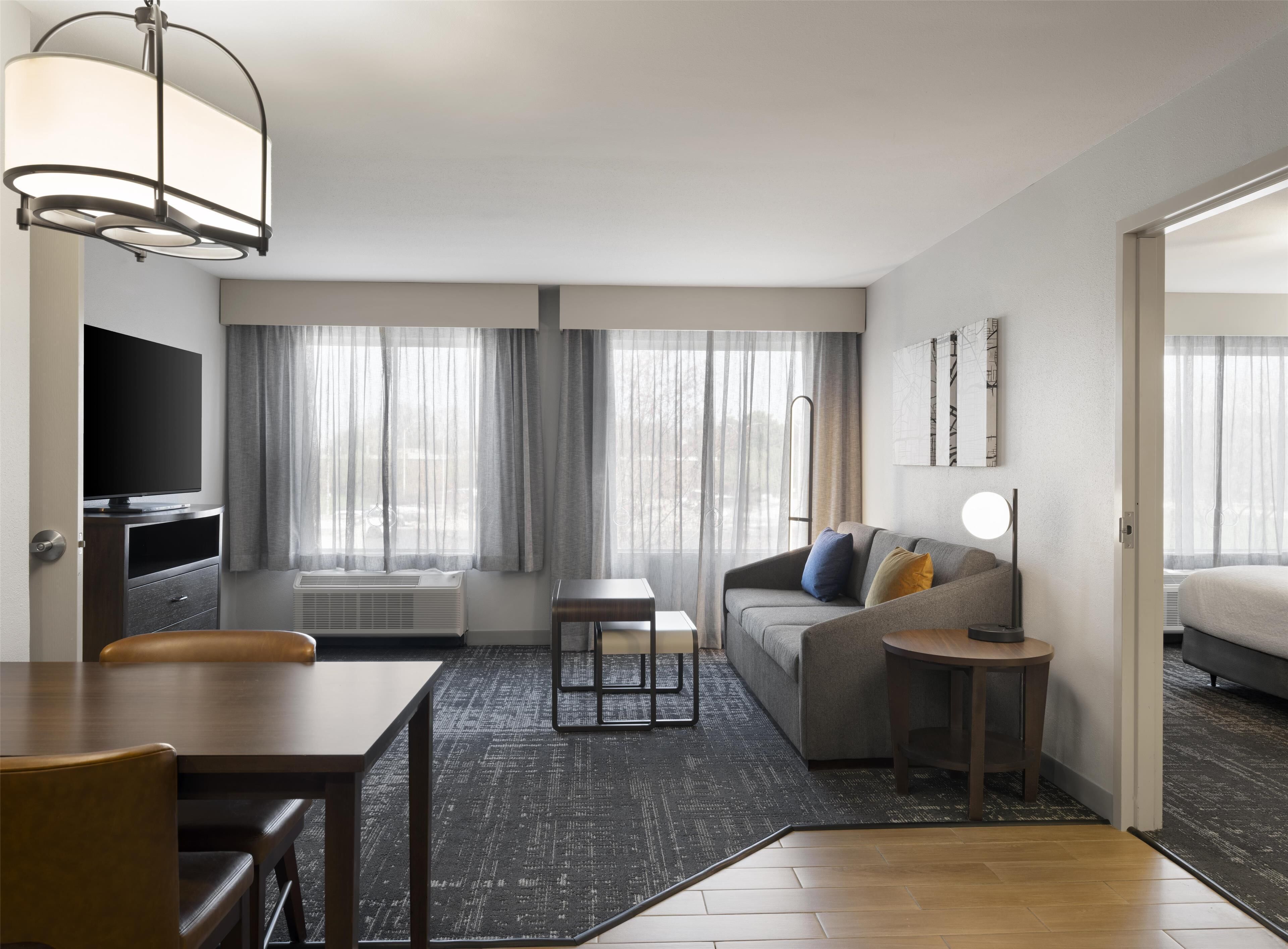 A modern hotel room with a sofa, a dining table, a TV, and a large window with curtains.