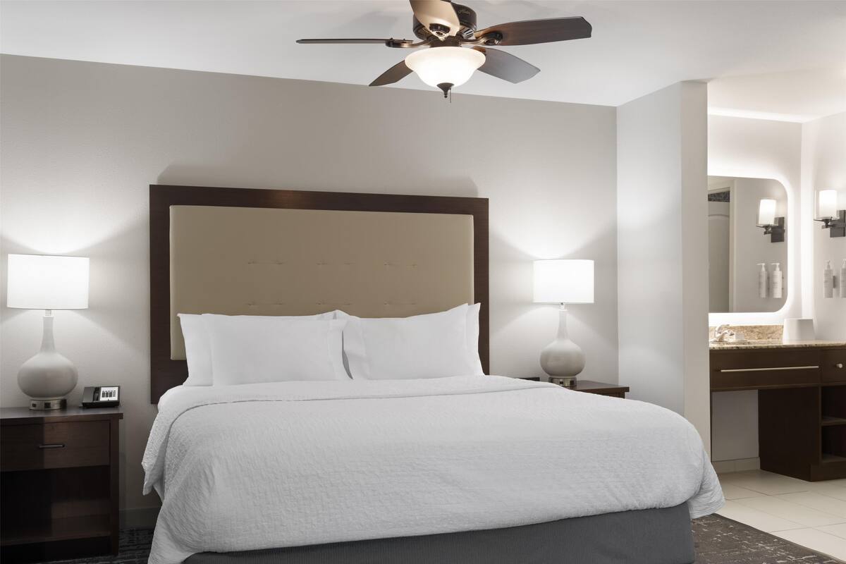 Ein Hotelzimmer mit einem großen Bett, zwei Nachttischen mit Lampen, einem Deckenventilator und einem Badezimmer mit Waschtisch und Spiegel.