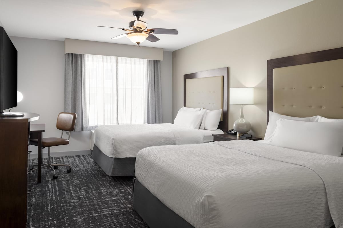 Ein Hotelzimmer mit zwei Betten, einem Schreibtisch, einem Stuhl, einem Deckenventilator und einem großen Fenster mit Vorhängen.