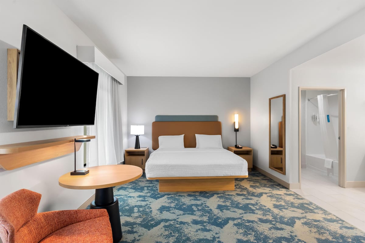 A hotel room with a large bed, a flat-screen TV mounted on the wall, a round wooden table, an orange chair, and a mirror on the wall.