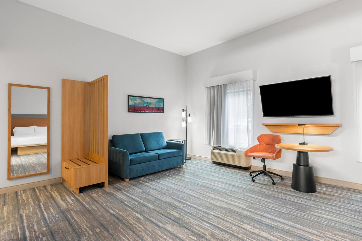 A hotel room with a blue sofa, a wooden wardrobe, a flat-screen TV, a round table, and a chair.