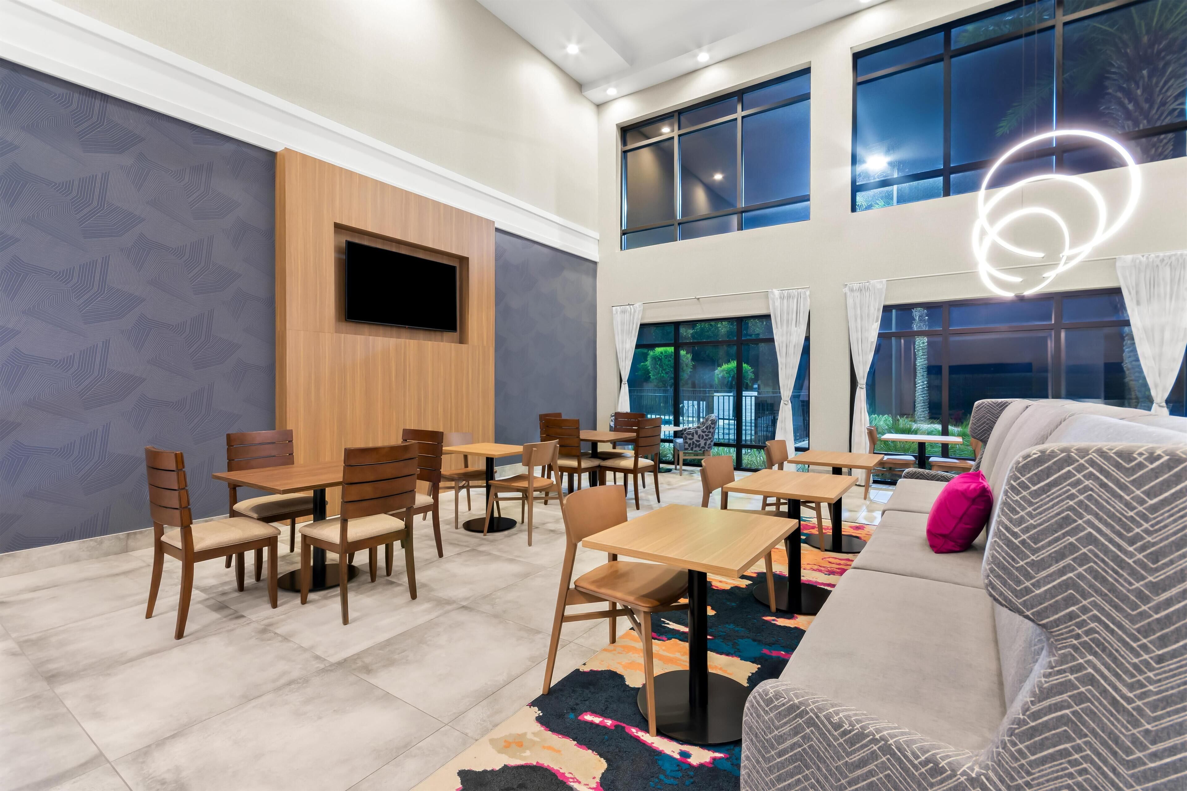 Un comedor moderno con mesas y sillas de madera, un sofá y ventanas grandes con cortinas.