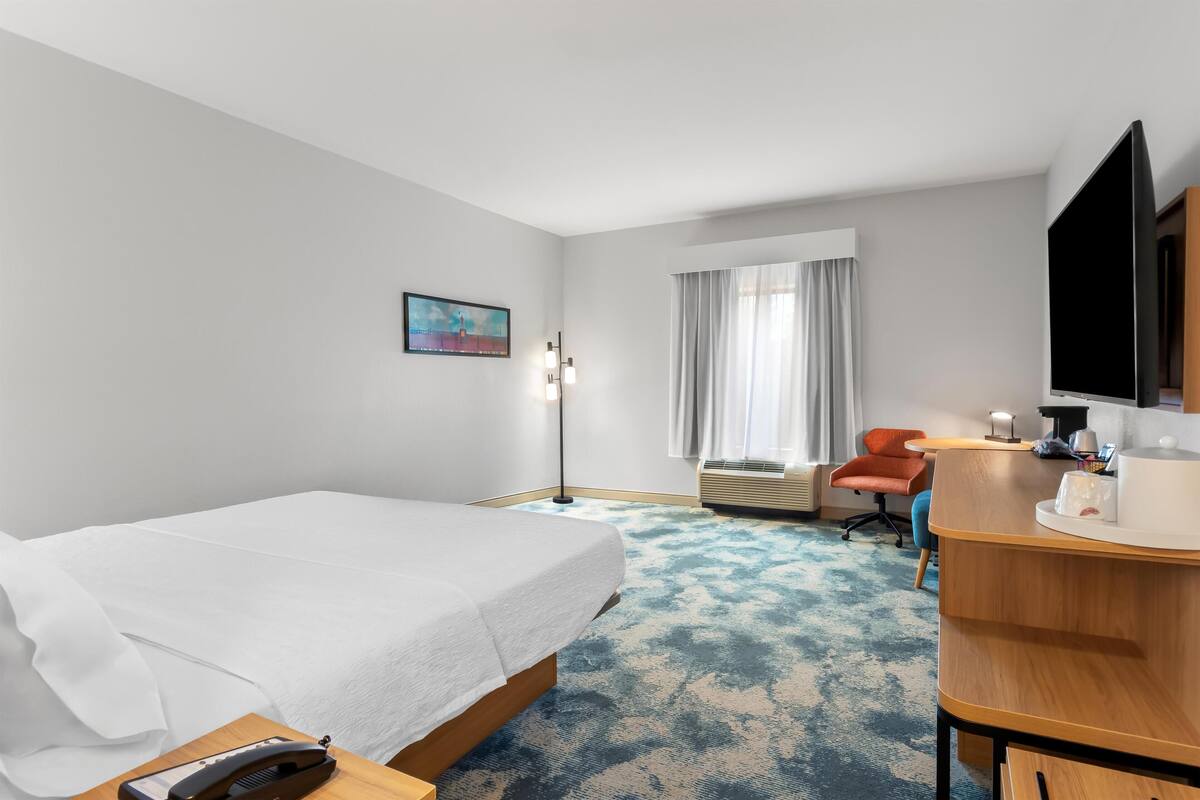 A hotel room with a bed, a desk, a chair, a flat-screen TV, and a window with curtains.