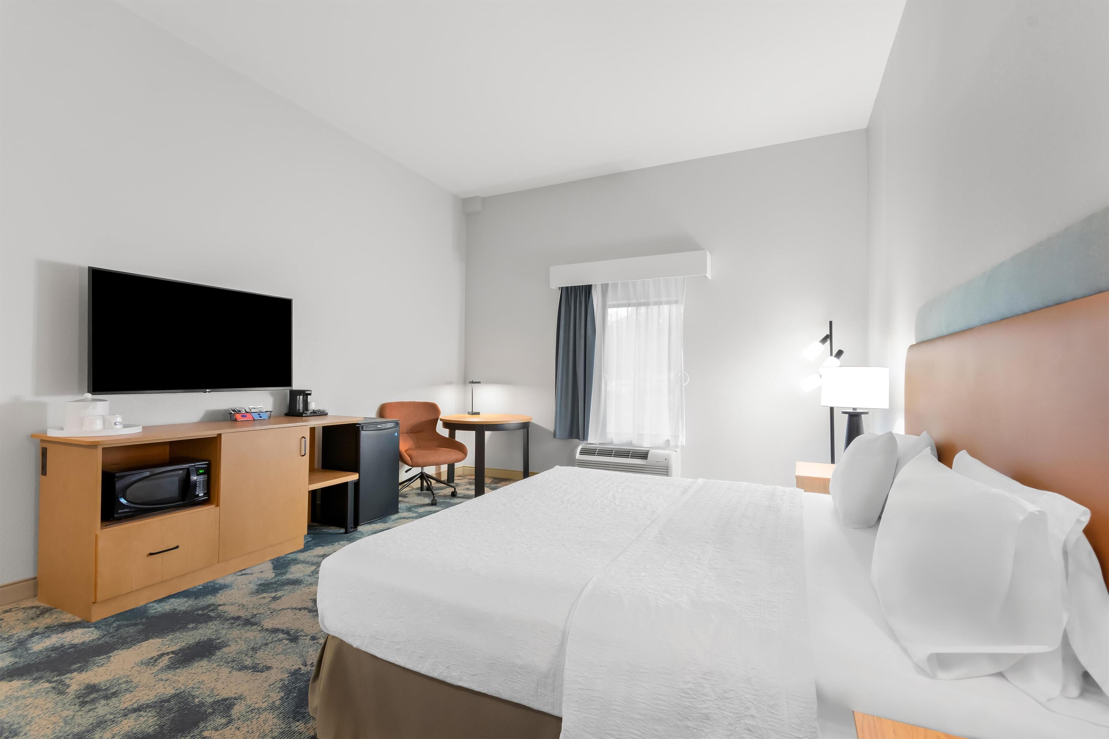 Une chambre d’hôtel comprenant un lit, un bureau avec un téléviseur à écran plat, un four à micro-ondes, une chaise, une petite table et une fenêtre avec des rideaux.