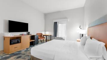 Une chambre d’hôtel comprenant un lit, un bureau avec un téléviseur à écran plat, un four à micro-ondes, une chaise, une petite table et une fenêtre avec des rideaux.