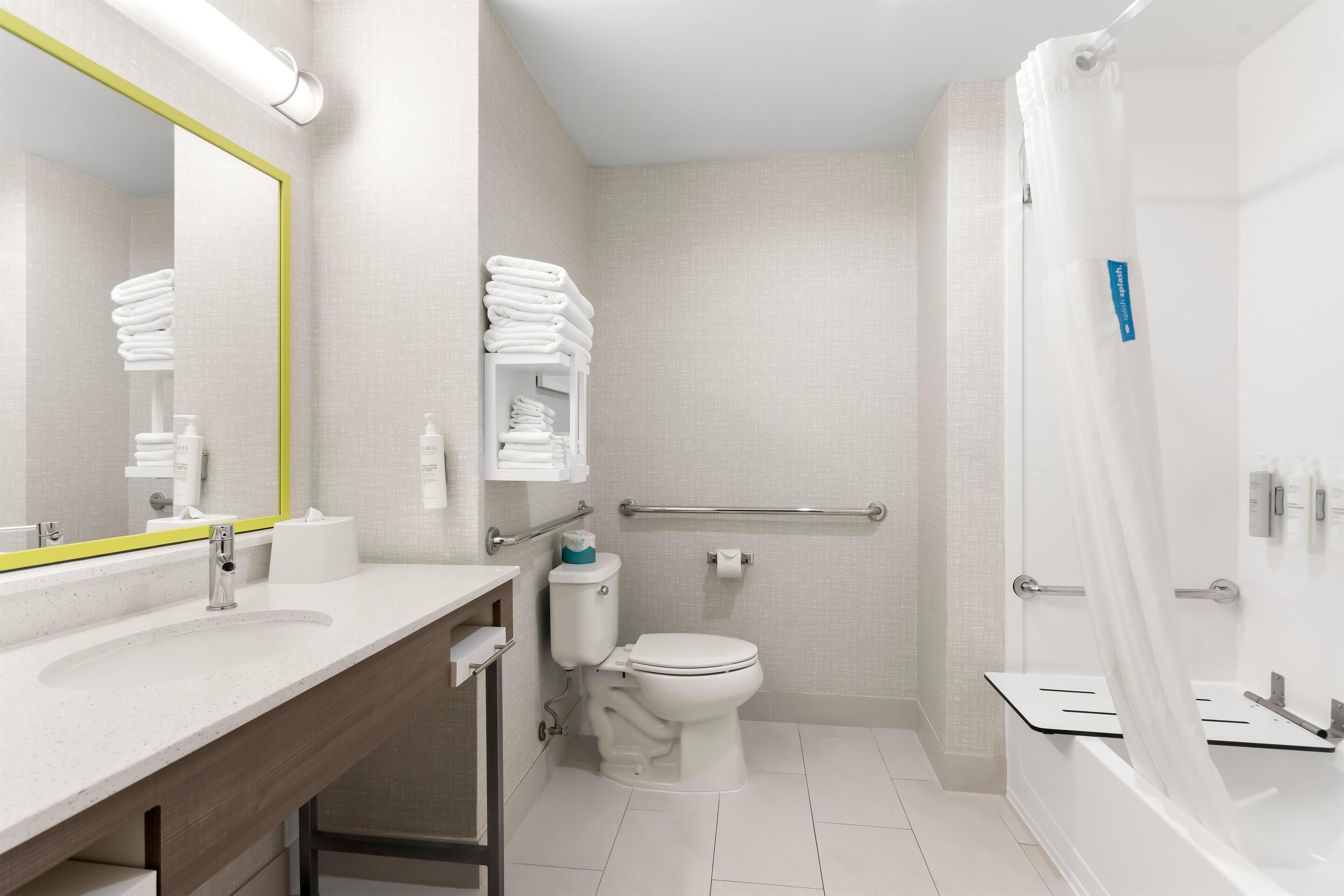 Une salle de bain moderne avec un lavabo, des toilettes et une baignoire, tous de couleur blanche. On peut voir des serviettes pliées sur une étagère, ainsi qu'une barre d'appui.
