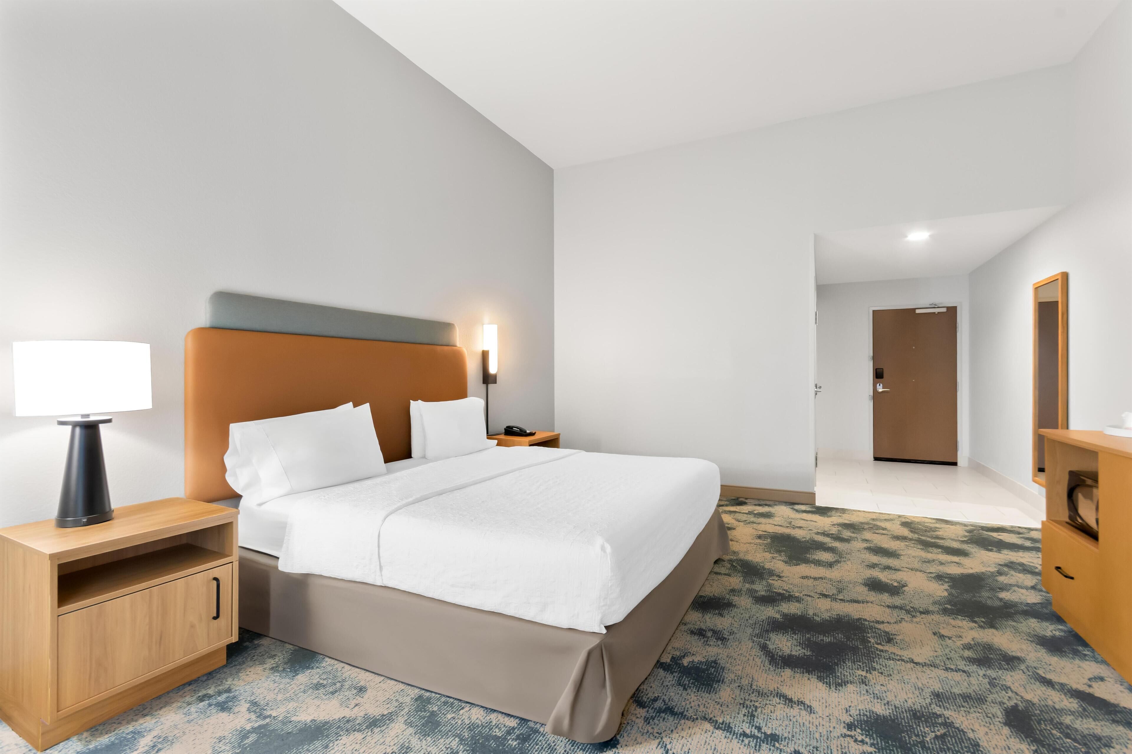 Habitación de hotel con cama, mesita de noche, lámpara, cómoda de madera y una puerta que da a otra habitación.
