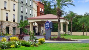 Een hotel met een heldere ingang, een bord met Hampton Inn & Suites by Hilton en een aangelegde omgeving met bloemen en palmbomen.