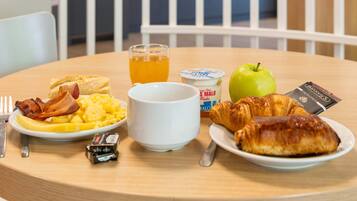 A breakfast table with scrambled eggs, bacon, toast, a croissant, an apple, a glass of orange juice, a yogurt cup, and a chocolate bar.
