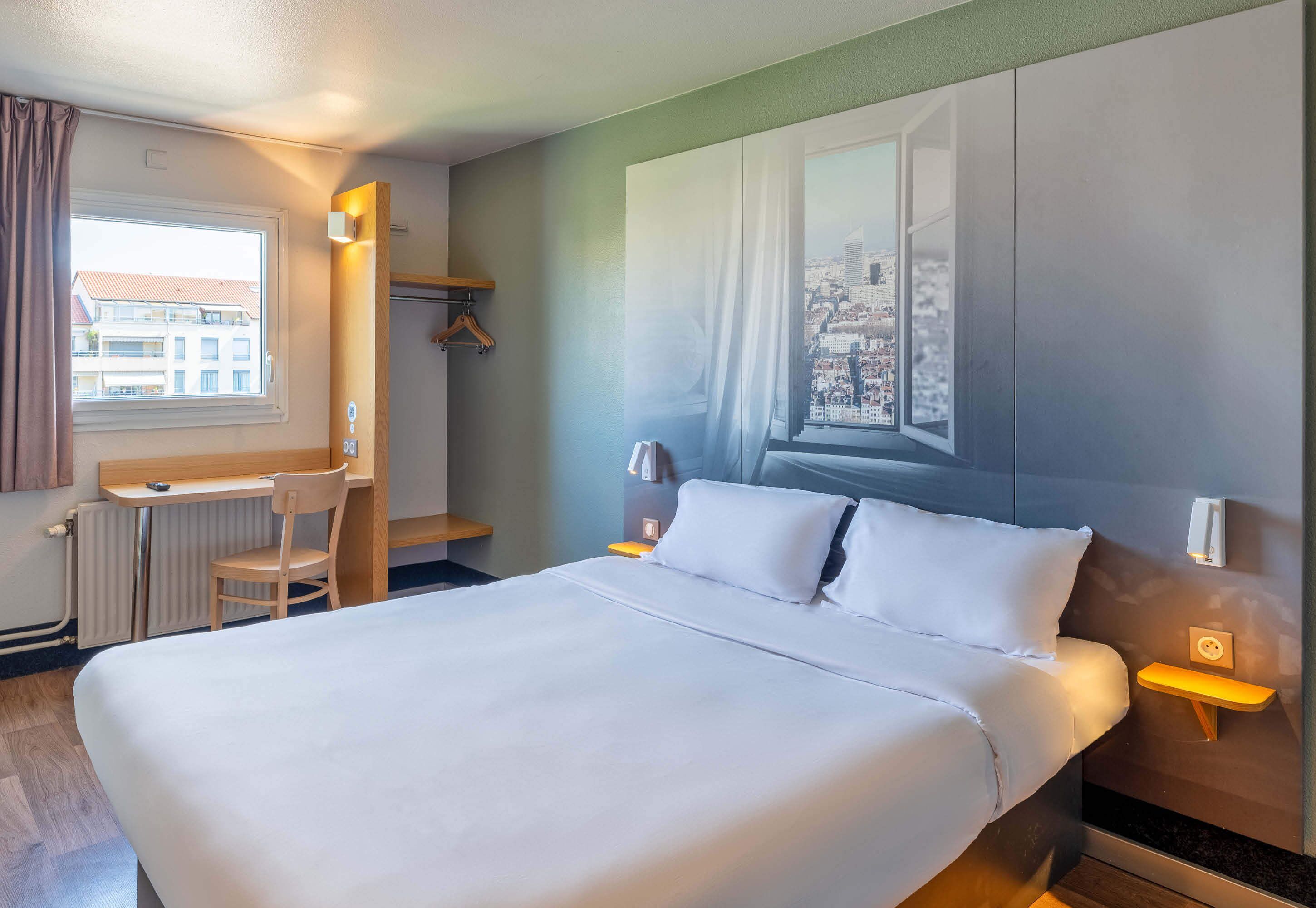 A hotel room with a bed, a desk, a chair, a window with a view of buildings, and a wall-mounted shelf.